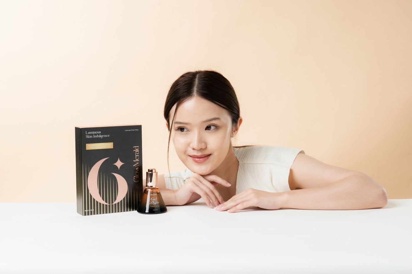 Model resting beside a GlowMerald sheet mask box and serum